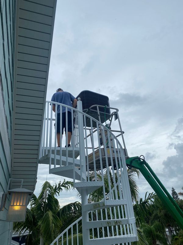 Man and object on a spiral staircase raised by a boom lift. Blue sky and foliage in the background.