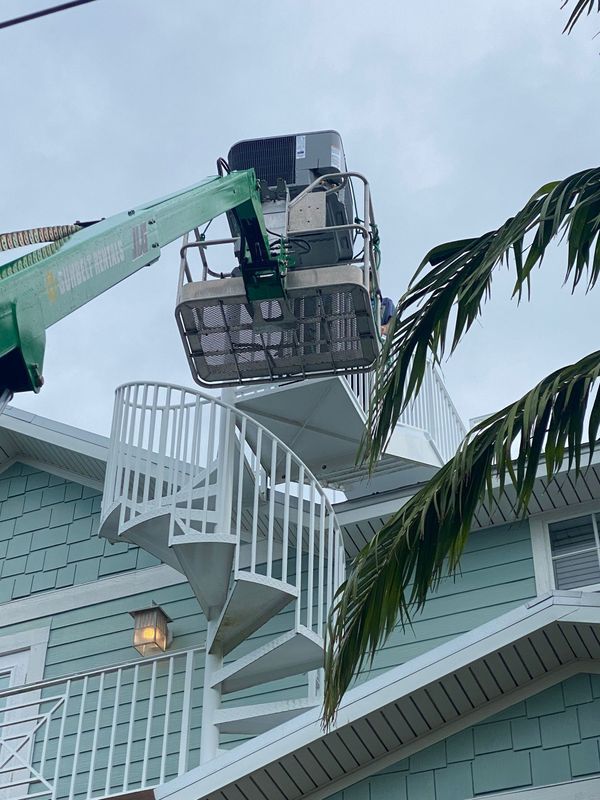 A lift raises an AC unit toward a building's roof next to a spiral staircase. Green lift arm, cloudy sky.