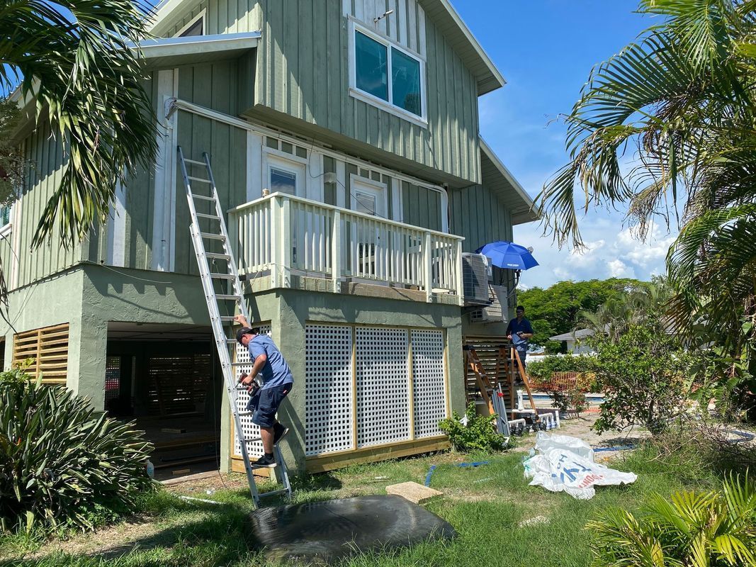 Two-story green house with a person on a ladder;  a person under a blue umbrella on the deck.