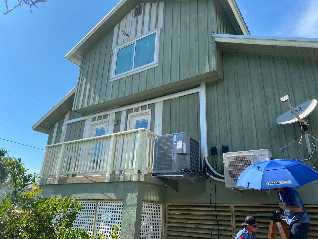 Two-story green house with balcony, AC units. Person on ladder with umbrella in front.