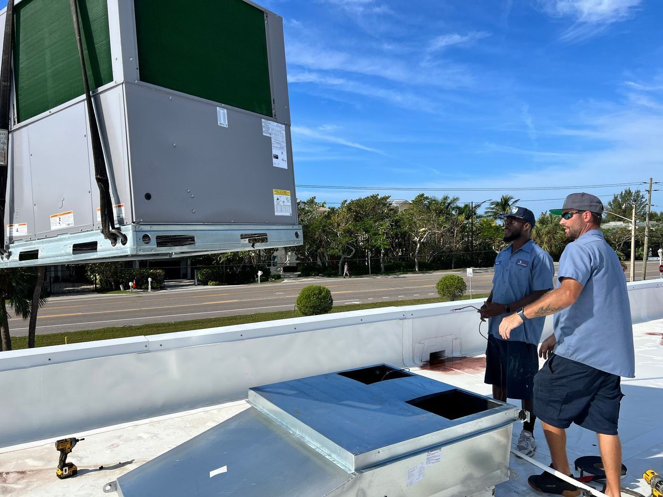 Two HVAC technicians on a rooftop watch a rooftop unit being lifted by a crane against a blue sky.