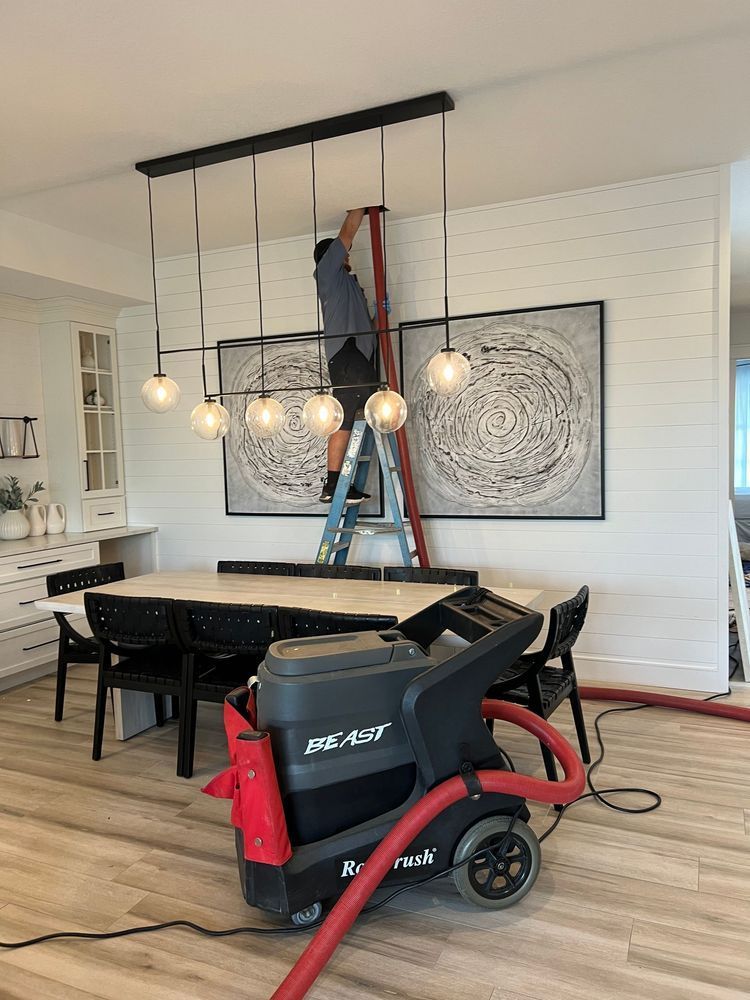 Person on a ladder working on a light fixture above a dining table with cleaning equipment nearby.