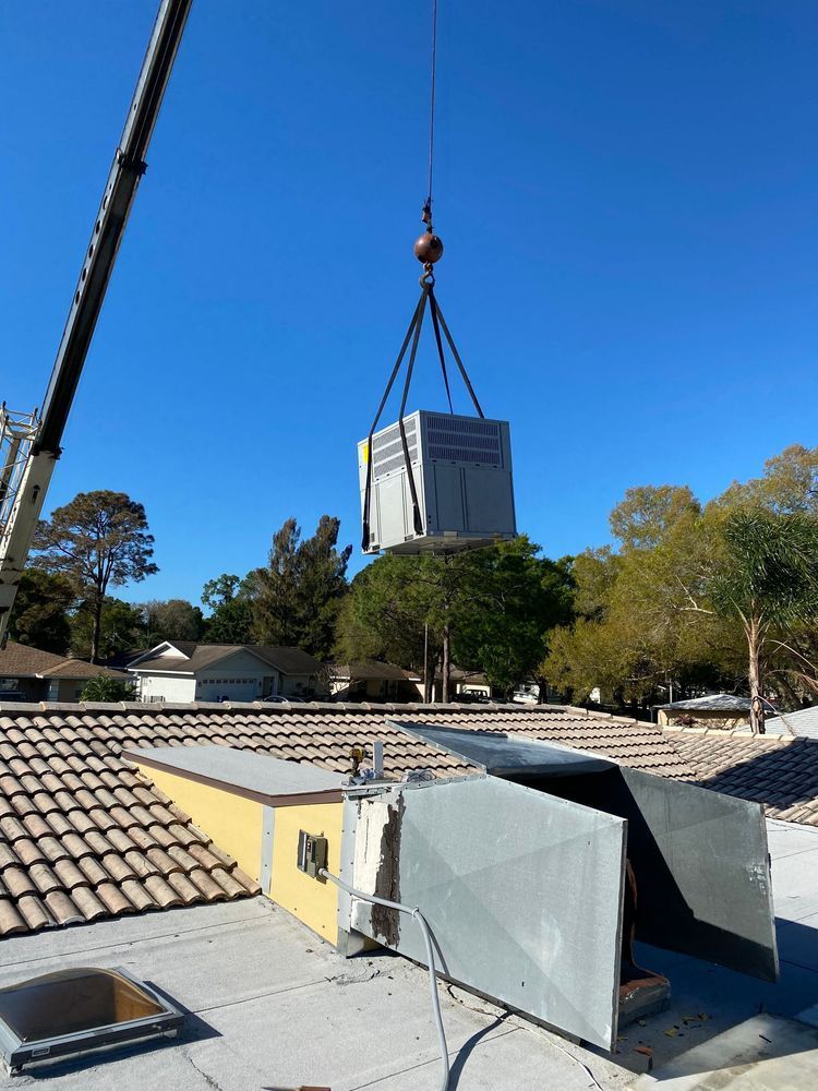 A crane lifting a large HVAC unit onto a rooftop. Bright blue sky, trees, and houses visible.