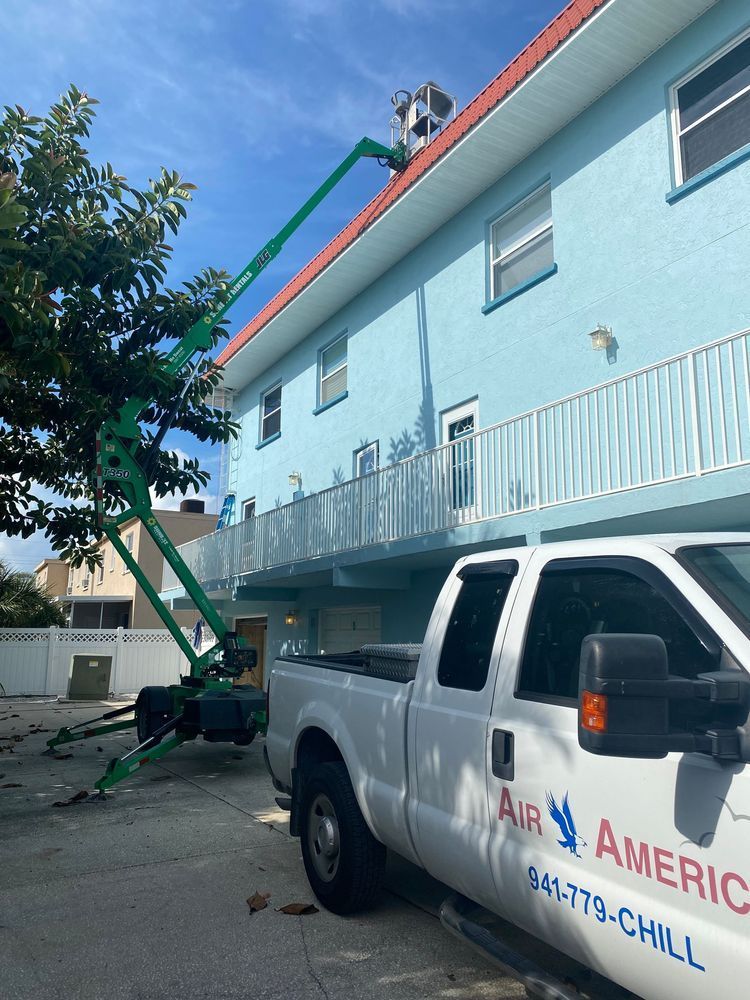 A green lift truck extends towards a light blue building's roof; a white truck is parked below.