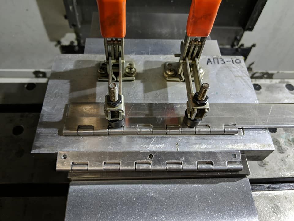 Metal hinges clamped to a metal fixture on a machine table with orange clamps.