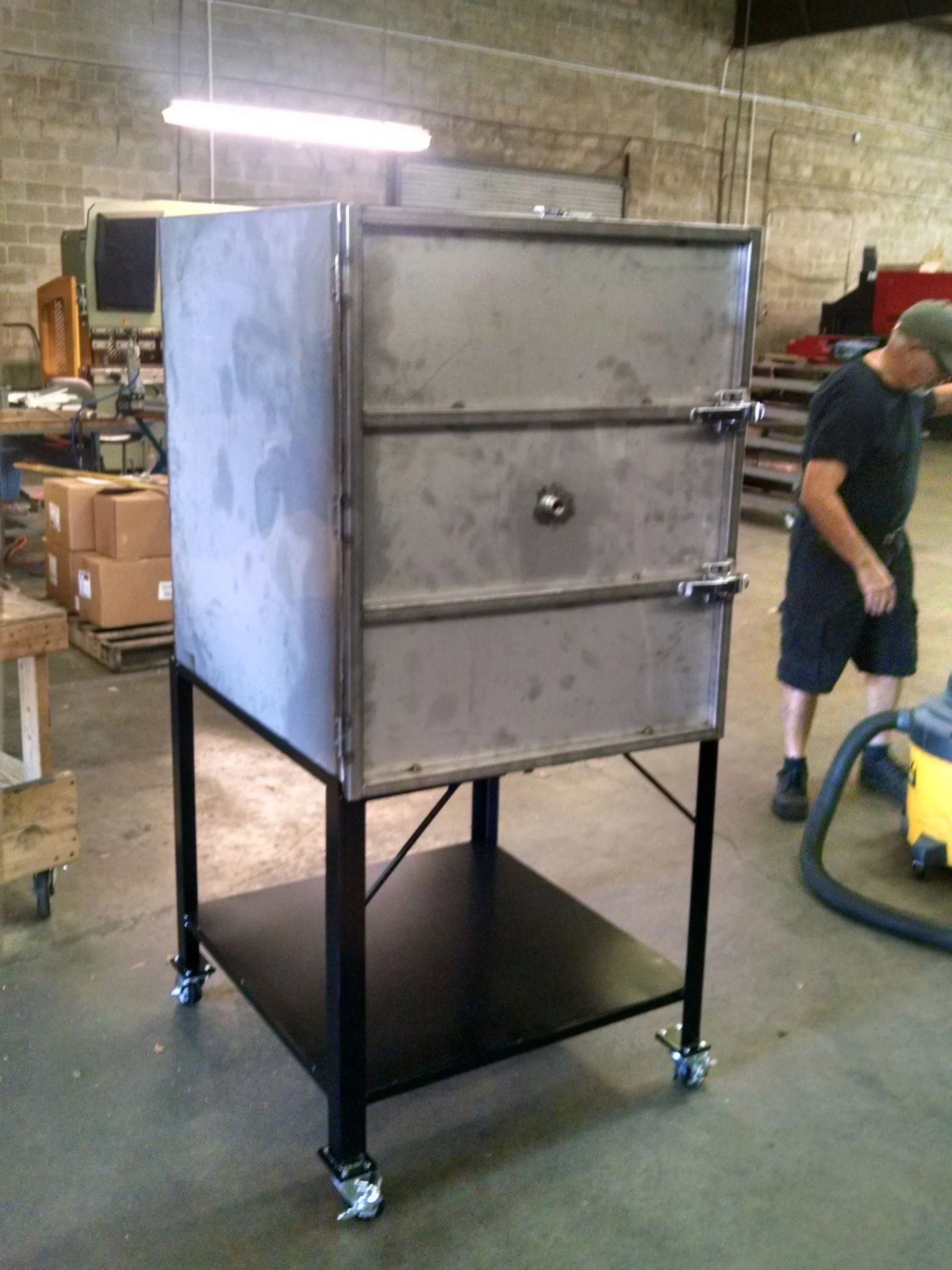 Large metal smoker on rolling cart with person near it in a workshop setting.
