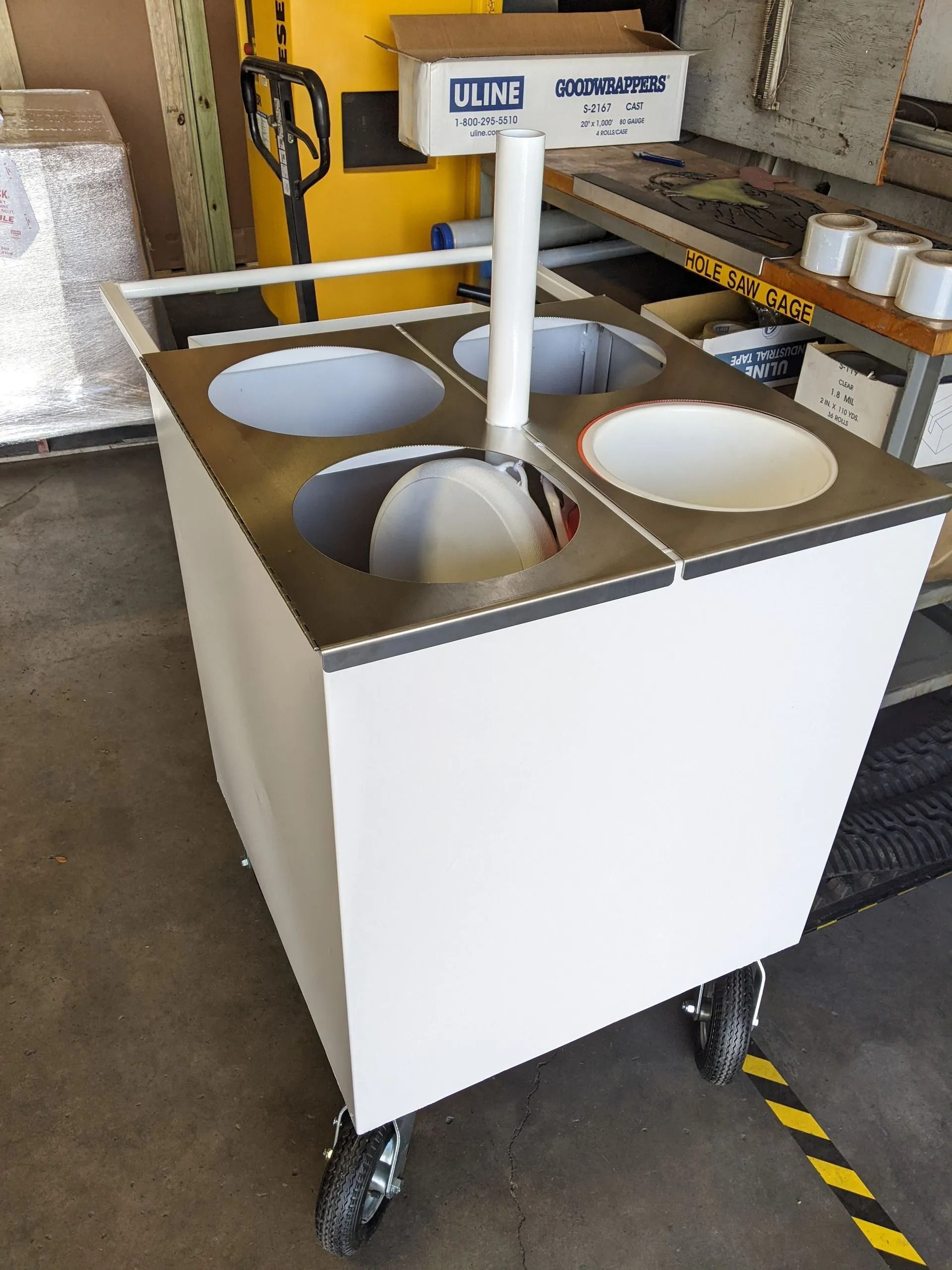 White cart with four inset bowls, a central pole, and casters. In an industrial setting.