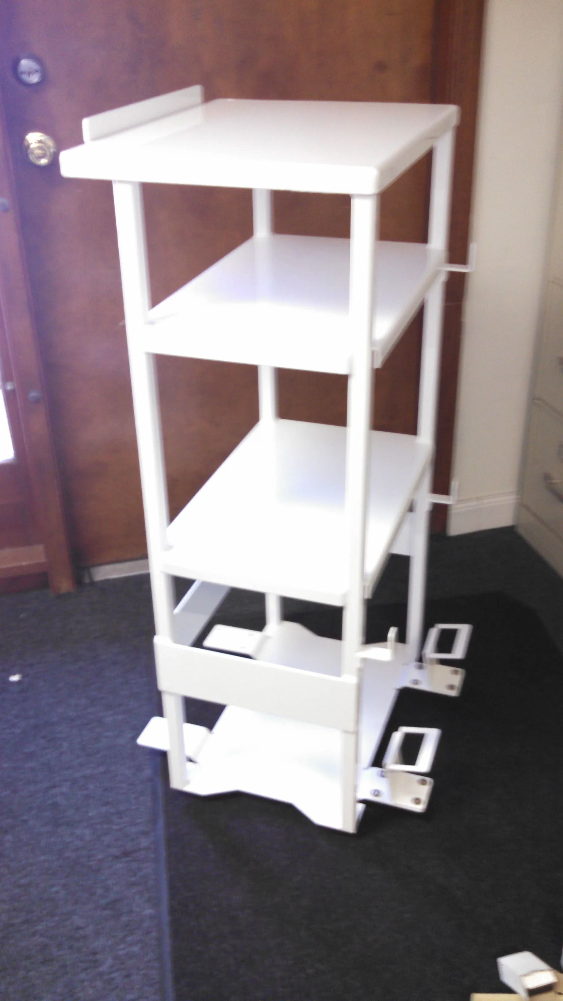 White multi-shelf cart on a dark rug next to a brown door and a white filing cabinet.