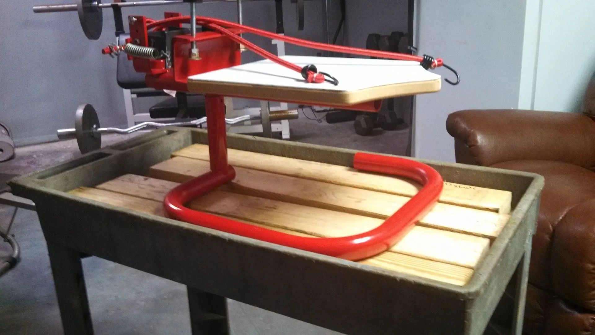 Red weightlifting apparatus on a wooden platform, with resistance bands attached. Set in a home gym.