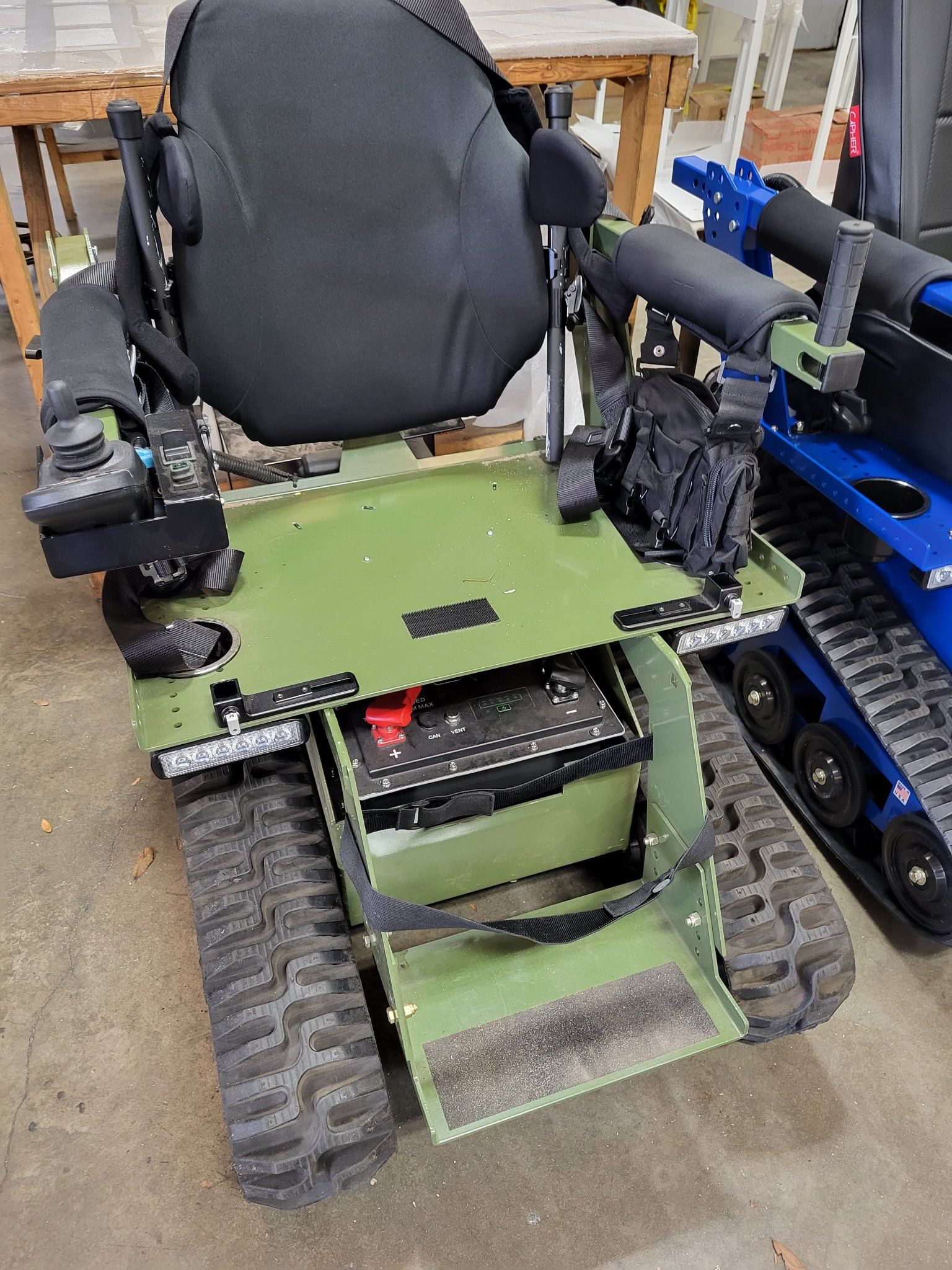 Green tracked wheelchair with black seat and armrests, inside a workshop.