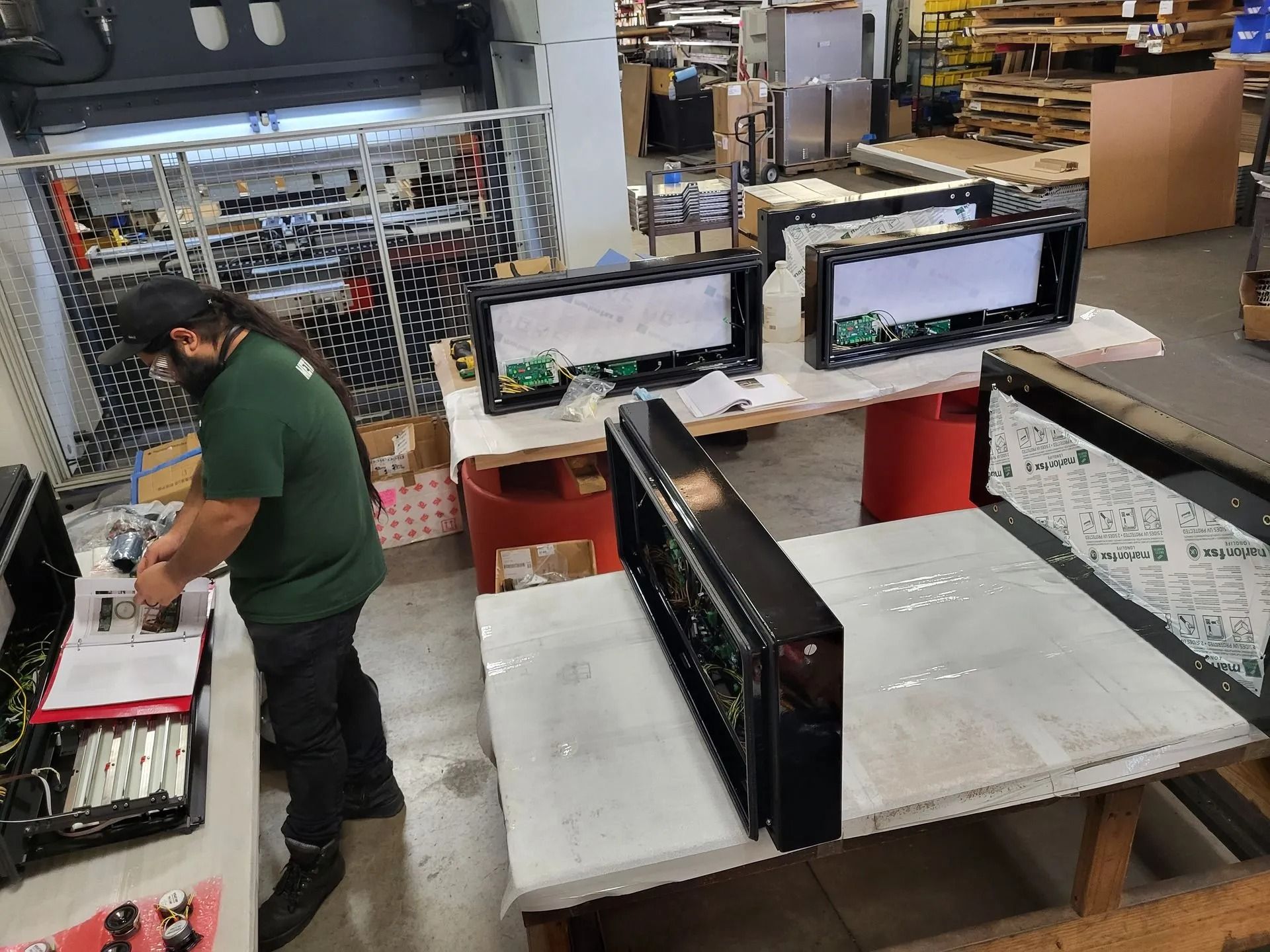 Man assembling electronic components at a workbench in a factory, with partially built displays on tables.