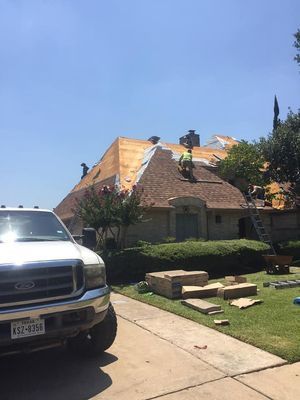Roofers installing shingles on a house with a large, sloped roof under a bright blue sky. A white truck is parked in the foreground.