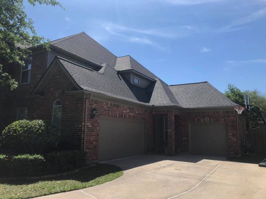 Two-story brick house with a dark gray roof, two-car garage, and green lawn, set against a blue sky.
