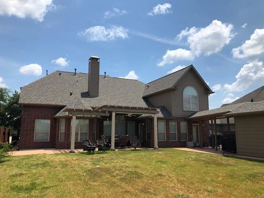 Backyard view of a red brick house with a brown roof on a sunny day. There is a patio with seating and a large grassy yard.