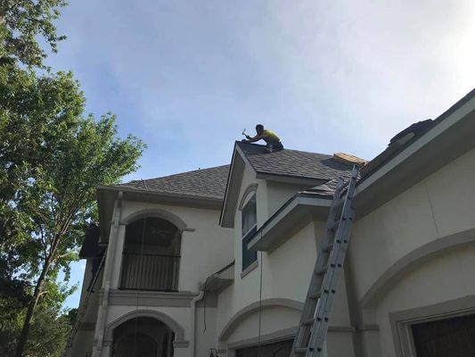 A roofer works on a light-colored house's roof under a sunny sky. A ladder leans against the side.