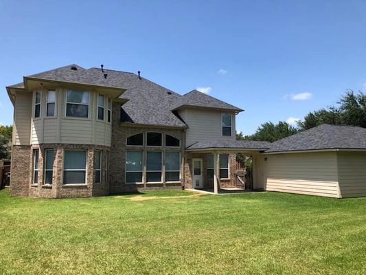 Back view of a two-story beige house with a dark gray roof and a grassy lawn under a blue sky.