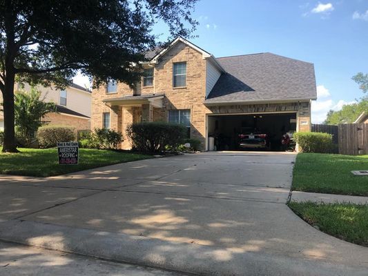 Two-story brick house with a driveway and garage, trees, and a lawn; sunny day. A car is parked in the garage.