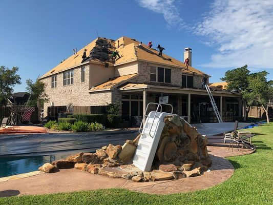 Roofers working on a two-story brick home, with a pool in the foreground. Blue sky and green grass.