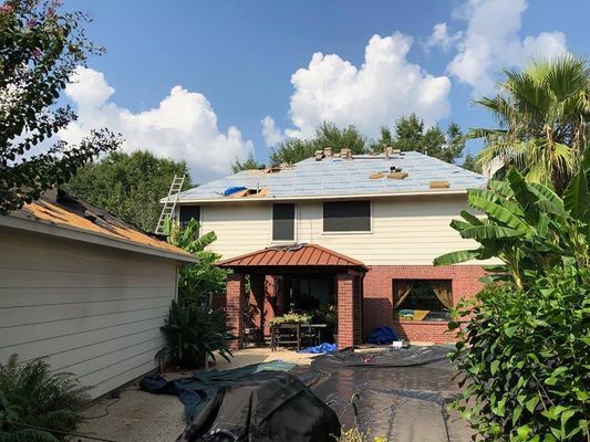 Two-story house under roof repair with materials and ladder visible. Blue sky with clouds, surrounded by trees and foliage.