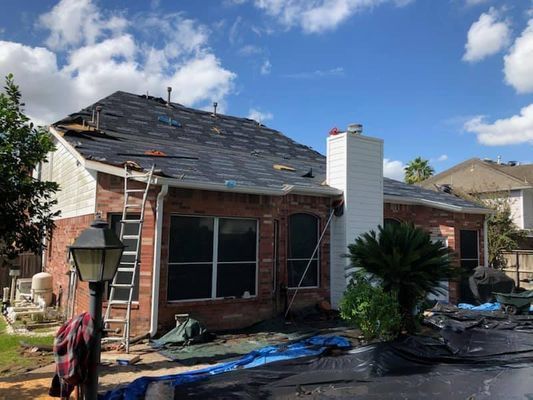 Roof being replaced on a brick house with a white chimney under a blue sky. A ladder leans against the house.