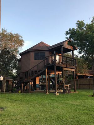 Two-story brown wooden structure with deck, accessed by exterior stairs, on a green lawn with trees and a fence in the background.