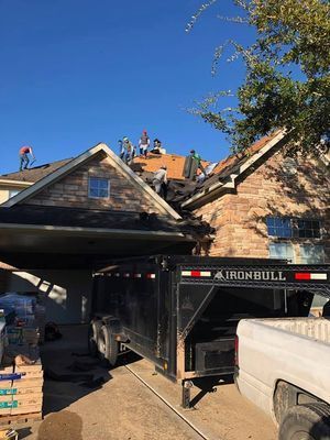 Roofers working on a house with a trailer and truck parked in the driveway on a sunny day.