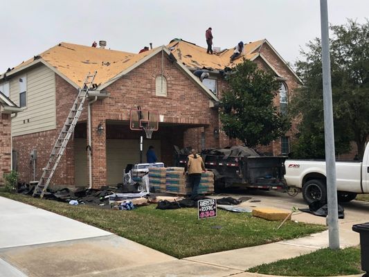 Roofers replace a shingle roof on a two-story brick house. Materials and a truck are in the driveway.