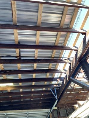 View from below a corrugated metal roof supported by wooden beams in an outdoor setting with blue sky visible.