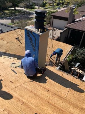 Two roofers working on a roof near a chimney, with a blue tarp and new wooden boards visible on the roof.