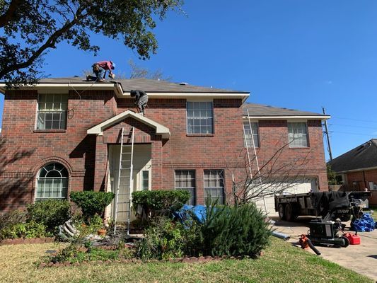 Two roofers working on a brick house's roof on a sunny day. Ladders are set up, and yard tools are visible.