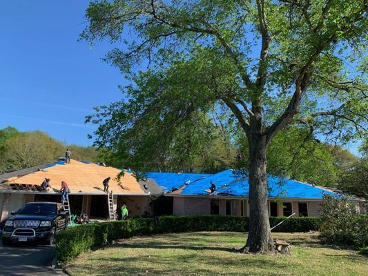 Roofers working on a house with some areas covered in blue tarps. The sky is blue, and there is a tree in the yard.