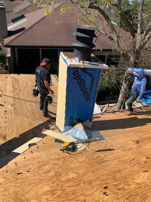 Two roofers working on a roof. One near a chimney with a blue wrap, the other near a tarp.