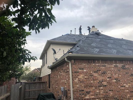 Workers on a partially covered roof, applying underlayment to a two-story house with brick and siding, cloudy sky.