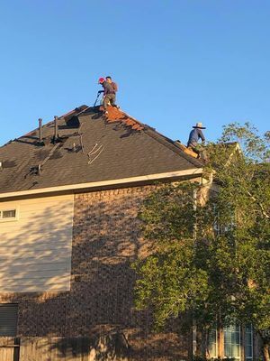 Two roofers repairing a shingled roof on a two-story brick home under a clear blue sky.