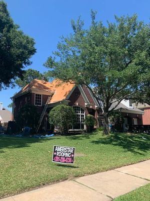 House undergoing roof replacement on a sunny day. A ladder leans against the partially torn-off roof, with a company sign in the front yard.