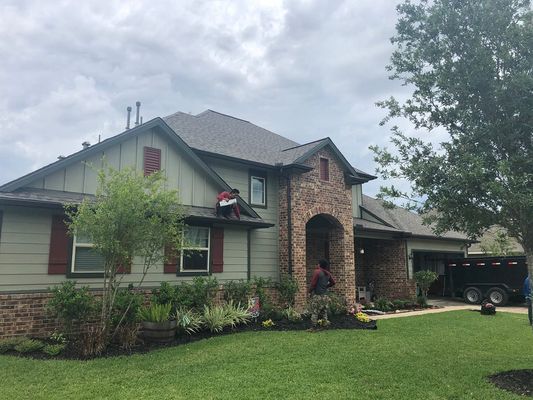 Two roofers working on a home with green siding, brick accents, and a dark gray roof. A trailer is in the driveway.