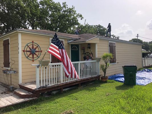 A yellow house with an American flag on the porch, and people on the roof. A green trash can sits in the yard.