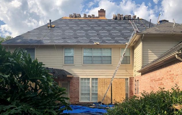 Roof being repaired on a two-story house. A ladder leans against the roof, and stacks of shingles sit on top.