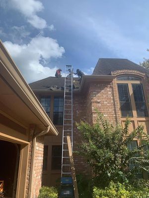 Two workers on a roof with a tall ladder, against a blue sky. A brick house and a garage are in the frame.