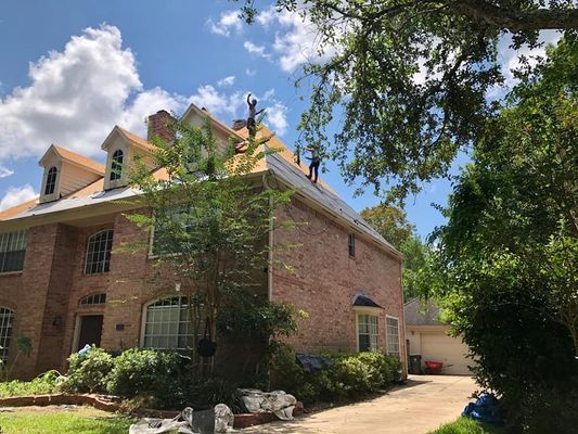 Two workers on a brick house roof on a sunny day. Green trees and a blue sky surround the home.