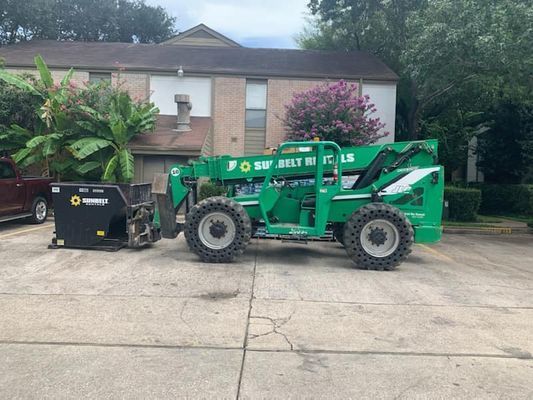 A green forklift with black attachments is parked on a concrete surface in front of a two-story apartment building.