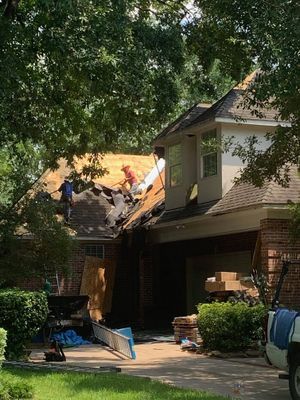 Workers removing old shingles from a house roof. Two workers stand on the roof, while another prepares materials below.