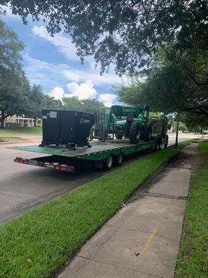A green flatbed trailer carrying a black container and a green forklift is parked on a street next to a sidewalk.