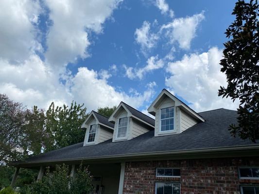 A house with three dormer windows against a cloudy blue sky. The roof is dark, and the house is brick.