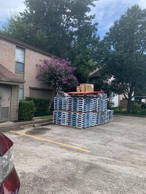 A stack of blue pallets in a parking lot next to a brick building and trees. Brown cardboard boxes rest on top of the pallets.