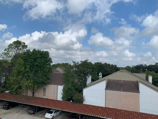 A sunny day with a partly cloudy sky over apartment buildings with a covered parking area. Green trees are visible.