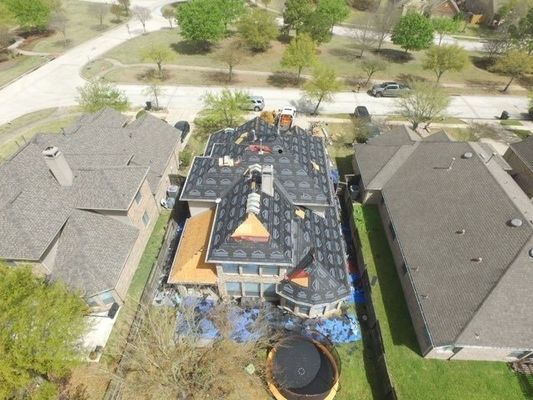 Aerial view of a house with roof under construction, showing exposed wood and new shingles, surrounded by other houses and trees.