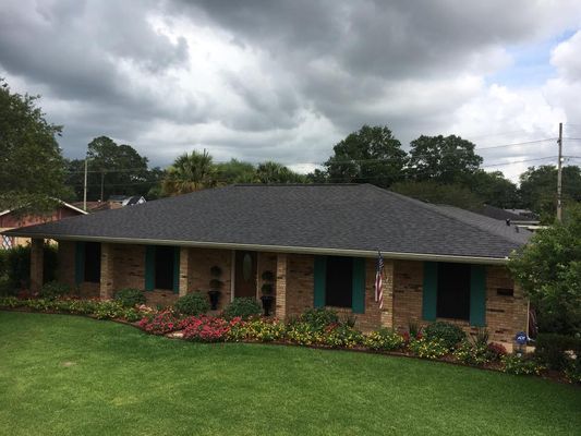 A one-story brick house with a dark gray roof, teal shutters, and a well-manicured lawn under a cloudy sky.