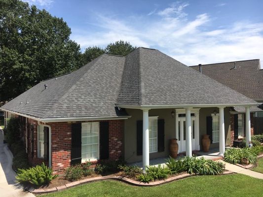 A brick and stucco house with a gray shingled roof and white columns supporting the front porch, set on a well-manicured lawn.