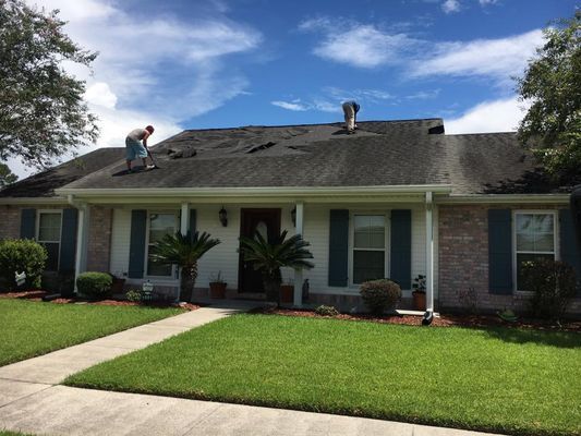 A person works on a damaged roof of a white house with blue shutters, under a partly cloudy sky.